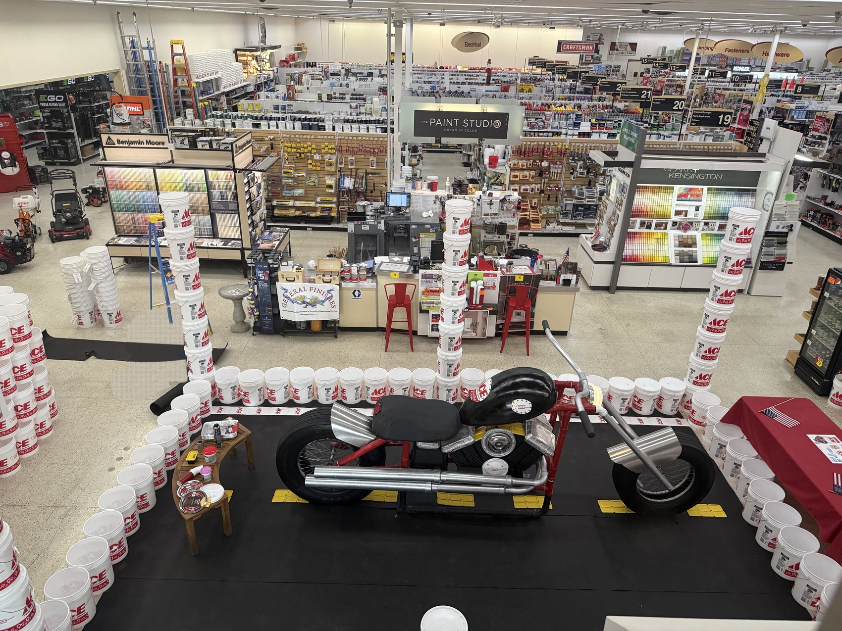 Looking down on the finished display from above: the full-size chopper stands on a black roadway mat with yellow centre-line markings, ringed by a corral of Ace buckets, with tall bucket columns rising on either side and the hardware store's aisles behind it.